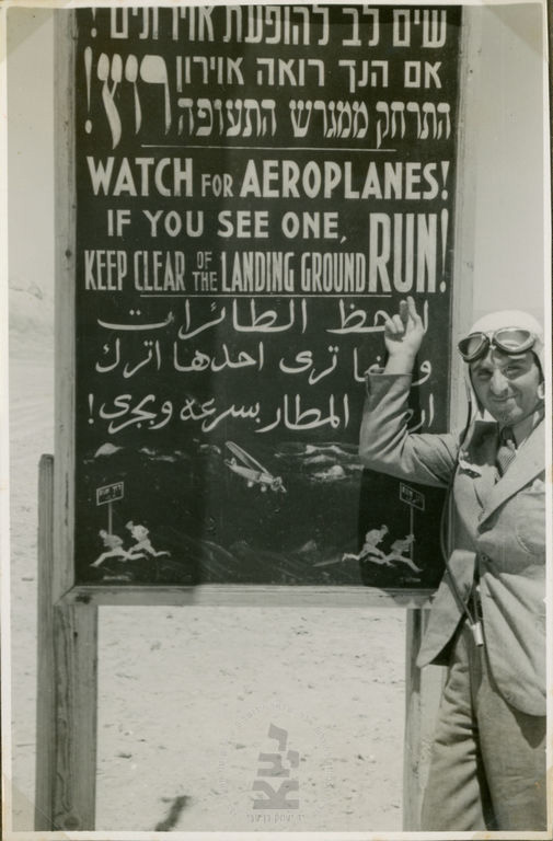 Zurr pointing to a sign in Hebrew, English, and Arabic (with a pictogram for the illiterate!) warning against moving aircraft at the Ashlag company airfield, Sodom, 1938–9 