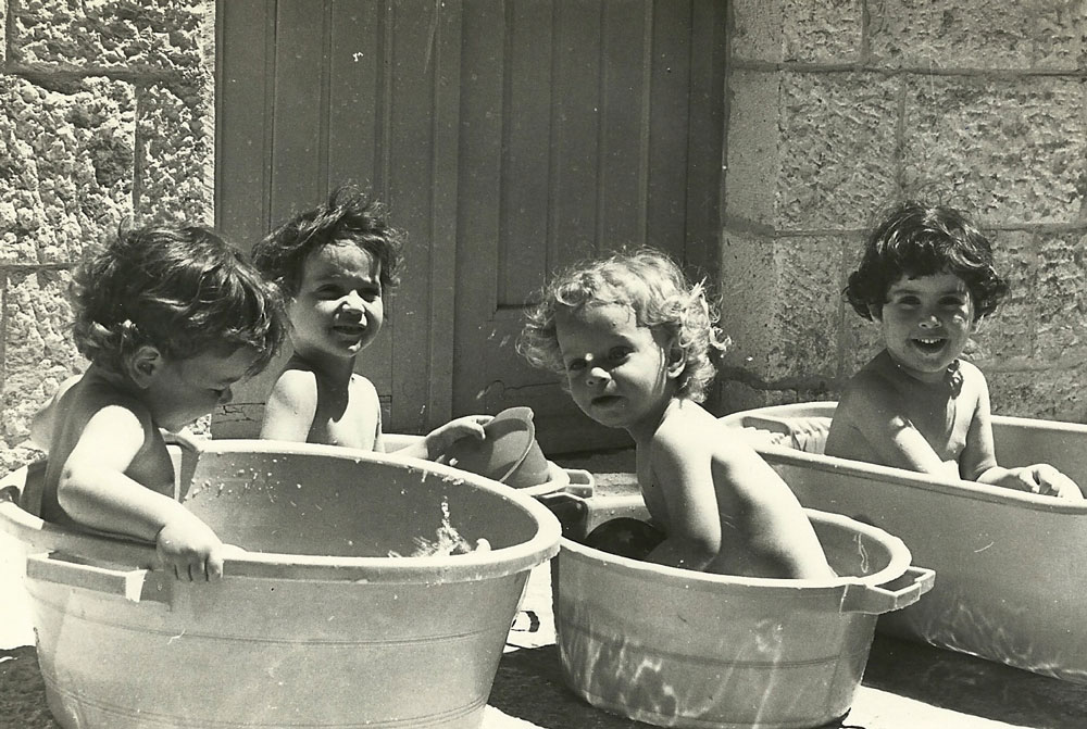 ילדים שוב משחקים וצוחקים בעברית ברחובות העיר העתיקה. ילדי הגן של גרעין גנ״ת מתרעננים בגיגיות מים ביום קיץ חם בחצר של התנחלות מוריה